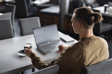 Freelancer man working by laptop PC sitting at table in a cafe drinking coffee, back view