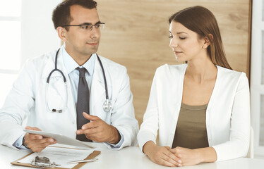 Doctor and patient discussing medical exam results while sitting at the desk in clinic. Male physician using tablet computer for filling up medication history record of young woman. Data in medicine