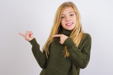 Wall Mural - Caucasian kid girl wearing green knitted sweater against white wall pointing and holding hand showing adverts