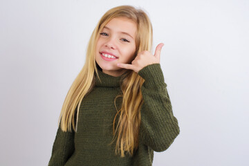 Wall Mural - Caucasian kid girl wearing green knitted sweater against white wall smiling doing phone gesture with hand and fingers like talking on the telephone. Communicating concepts.