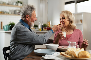 Wall Mural - Senior couple eating breakfast in the kitchen. Husband and wife talking and laughing while eating a sandwich.