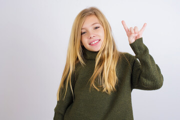 Wall Mural - Caucasian kid girl wearing green knitted sweater against white wall doing a rock gesture and smiling to the camera. Ready to go to her favorite band concert.