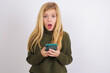 Shocked Caucasian kid girl wearing green knitted sweater against white wall opens mouth hold phone reading advert unbelievable big shopping prices
