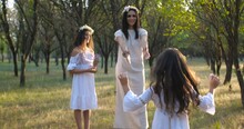 Tracking Shot  Mother Walking With Her Daughters Wearing Flower Wreaths And White Boho Dresses  On The Grass On A Nature Background. Mother’s Day Concept. 4k 50 Fps Slow Motion
