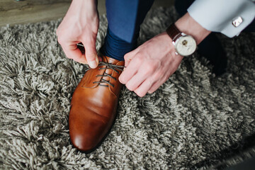 Wall Mural - Brown leather shoe lacing. Businessman in white shirt and suit trousers. Groom getting ready for the wedding. Wearing clothes background. Dressing up male fashion.