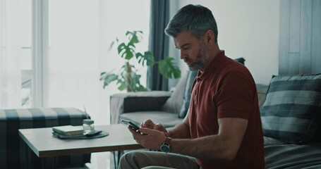 Wall Mural - Adult businessman sitting on the sofa using smartphone getting notification reading business news social media checking internet content at home.