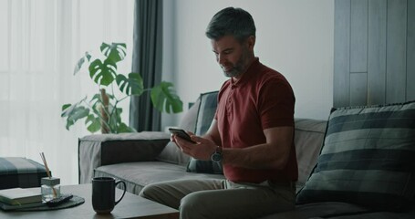 Wall Mural - Business adult mature handsome caucasian bearded man sitting on sofa using smartphone for typing messages. Lockdown and people. Casual home lifestyle.