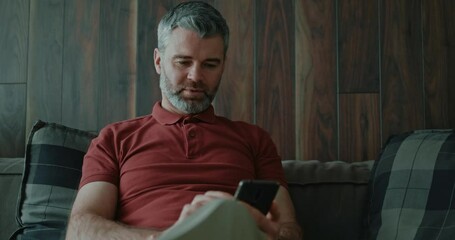 Wall Mural - Adult business male resting at home with social media application. Cheerful caucasian bearded man texting messages in mobile phone sitting on the sofa.