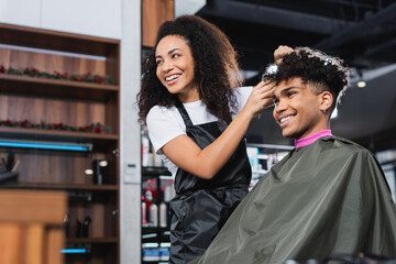 Cheerful african american hairstylist and client looking away in salon