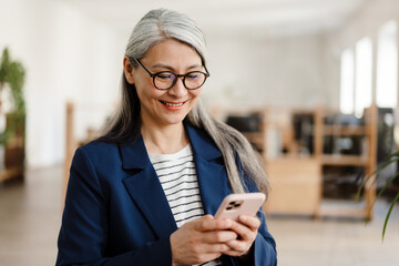 the smiling asian woman with glasses standing and looking at phone in office