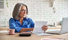 Adult Senior 60s Woman Working At Home At Laptop. Serious Middle Aged Woman At Table Holding Document Calculating Bank Loan Payments, Taxes, Fees, Retirement Finances Online With Computer Technologies