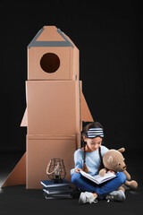 Poster - Cute little girl with cardboard rocket, books and toy on dark background