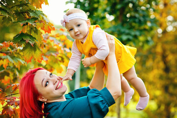 Little daughter and her mother in the autumn season in the park. A woman with red hair holds a baby in her arms against the background of an autumn maple branch.