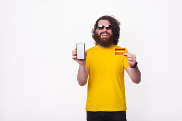 Cheerful bearded man in yellow t-shirt with long curly hair showing smartphone screen and credit card