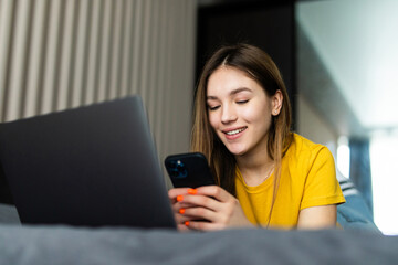 Portrait of beautiful young woman using her mobile phone in the bed with laptop.