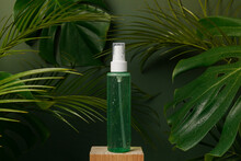 Cosmetic Green Bottle Of Skincare Product With Tonic Or Micelar Water On Green Background With Palm And Monstera Leaves. Natural Beauty Product With Water Drops