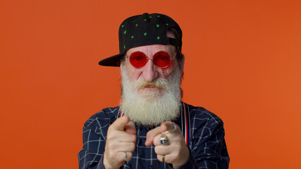 Portrait of seductive senior old bearded gray-haired man wearing sunglasses, charming smile, pointing at camera. Posing isolated on orange background. People emotions. Elderly grandfather pensioner