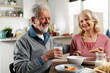 © JustLife - Senior couple eating breakfast in the kitchen. Husband and wife talking and laughing while eating a sandwich.