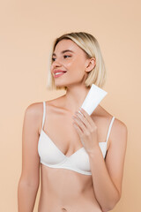 joyful woman in white bra looking away while holding cosmetic cream isolated on beige