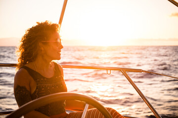 Wall Mural - Cheerful attractive young adult blonde woman smile and enjoy the excursion trip sitting on the sail boat deck looking the blue sea around - concept of summer adventure people