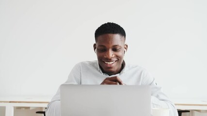 Sticker - A pleased black guy with earbuds is using a video connection on his laptop sitting alone in the office