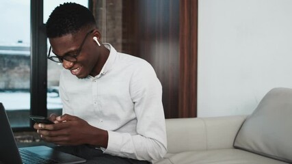 Poster - A smiling african american businessman wearing earbuds is using his cell phone while working with his laptop computer sitting on the sofa inside the apartments