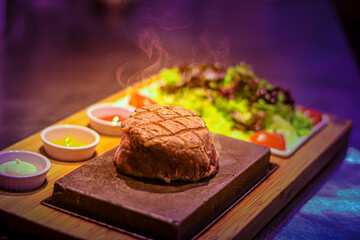 Poster - Grilled juicy beef steak smoking on hot stone served on wooden board with salad and sauces, colorful background