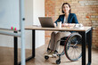 © Drobot Dean - White young woman in wheelchair working with laptop at office