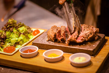 Poster - chef's hand cooking smoking steak on hot stone with sauces and garnishes on the side