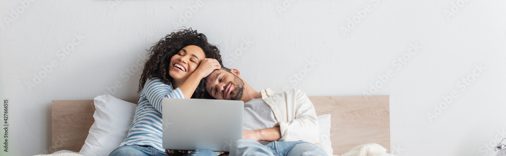 cheerful interracial couple resting in bed near laptop, banner - obrazy, fototapety, plakaty 