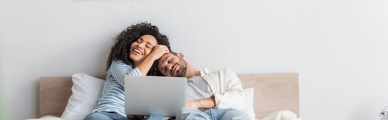 cheerful interracial couple resting in bed near laptop, banner