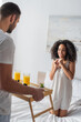 © LIGHTFIELD STUDIOS - blurred bearded man holding tray with breakfast near happy african american girlfriend
