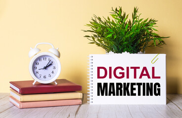 Fototapete - DIGITAL MARKETING is written in a notebook next to a green plant and a white alarm clock, which stands on colorful diaries.