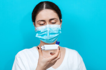 Bronchial asthma. Blurry portrait of a woman in a medical mask holding an inhaler in front of her and looking it. Blue background. Copy space