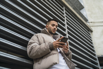 Wall Mural - Low angle shot of a sexy male in a street style outfit using his phone outdoors