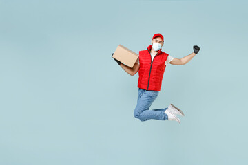 Wall Mural - Full length delivery guy employee man in red cap t-shirt vest uniform sterile face mask work courier service covid-19 flu virus hold empty cardboard box run go jump isolated on pastel blue background.