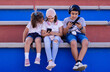 © Brian Díaz - Three Caucasian children sit together on bleachers, laughing as they look at their smartphones.