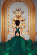 A young girl in a green haute couture feather dress standing with her back in a luxurious gold palace interior.