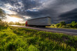 Motion blurred red truck driving on the asphalt road in rural landscape at sunset with dark storm cloud