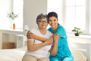 Portrait of supportive carer with her elderly patient who has Alzheimer. Happy retired senior woman together with caring young nurse or caregiver hugging and smiling sitting on bed in retirement home