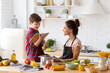 © deniskomarov - Happily smiling mother and son preparing dinner on loft kitchen. Boy sitting on table reading recipe from cookbook to cheerful mom wearing apron. Happy family weekend. Excited housework