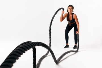 Sporty young woman in black suit doing crossfit exercises with ropes over isolated white background. hard workout. wave