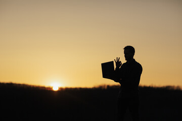 Man talking on video communication and shows ok sign. Man works behind a laptop on the background of orange sunset. Copy space.