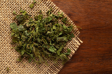 Wall Mural - Macro close-up of Organic green dry Fenugreek leaves (Trigonella foenum-graecum) on the wooden top background and jute mat. Pile of Indian Aromatic Spice. Top view