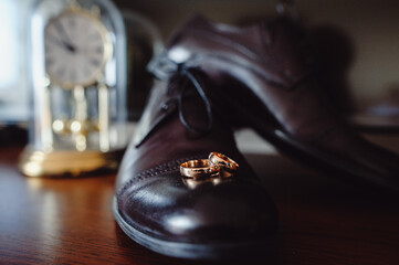 Wall Mural - Stylish men's black shoes, two wedding rings lying on a brown table. Wedding accessories bride on wooden background. composition of idea. holiday concept. Selective focus. Top view.