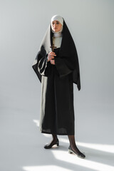 Wall Mural - full length view of young nun in black vestment standing with crucifix on grey background