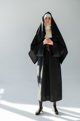 Wall Mural - full length view of nun with crucifix looking at camera on grey background with shadows