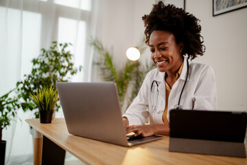 Afro American female doctor in uniform and stethoscope working on laptop in modern hospital.