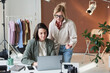 © AnnaStills - Young designer sitting at the table with laptop and discussing online work together with her colleague at studio