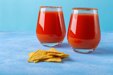 Traditional Mexican cuisine: Nachos, corn tortilla chips with spicy sauce and alcoholic drink Michelada. Two glass of Cocktail with tomato juice and beer on blue background. Flat lay, close up